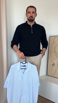 Schwarzer Reißverschluss-Pullover über beigen Hosen, der ein hellblau gestreiftes kurzärmliges Hemd an einem Haken hält.