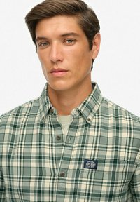 Zielono-biała koszula w kratę zapinana na guziki, wykonana z teksturowanej tkaniny, z naszywką z logo oraz brązowymi guzikami na kołnierzyku i mankietach.