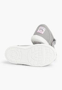 Zapatilla de lona gris con suela de goma blanca, que presenta una correa y una etiqueta colorida con un diseño de cartoon, etiquetada como "Hecho en España".