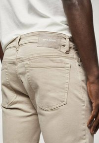 Gros plan d'une personne portant un jean beige MNG Denim et une chemise blanche, montrant les poches arrière et le bras droit reposant le long du corps.