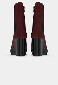 Stivali alla caviglia bordeaux con tomaia in suede, pannelli laterali elasticizzati neri e tacco spesso nero. Dotati di una zip posteriore per la chiusura.