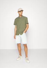 Chemise verte olive à manches courtes avec un col montant, fermeture à boutons ; associée à un short en denim bleu clair et des baskets crème. Casquette beige incluse.