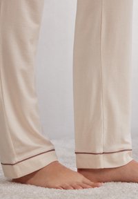 Beige pyjamabroek gemaakt van zachte stof, met een recht model, voorzien van contrasterende bruine stiksels bij de boorden. Geen zichtbare patronen.