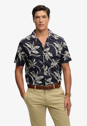 Camisa de manga corta azul marino con estampado de hojas blancas, con cuello de solapa y cierre de botones, combinada con pantalones kaki claro.