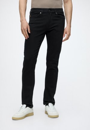 Homme portant un jean slim noir et des baskets blanches, debout devant un fond clair uni.