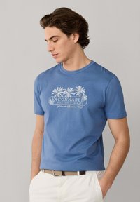 Junger Mann trägt ein blaues Façonnable-T-Shirt mit Palmenmotiven, weiße Hose und einen beigen geflochtenen Gürtel, Hände in den Taschen, blickt nach unten.