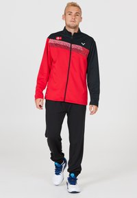 Rød og sort zip-up jakke med et horisontalt stribedesign, båret med sorte bukser og hvide sko med blå detaljer.