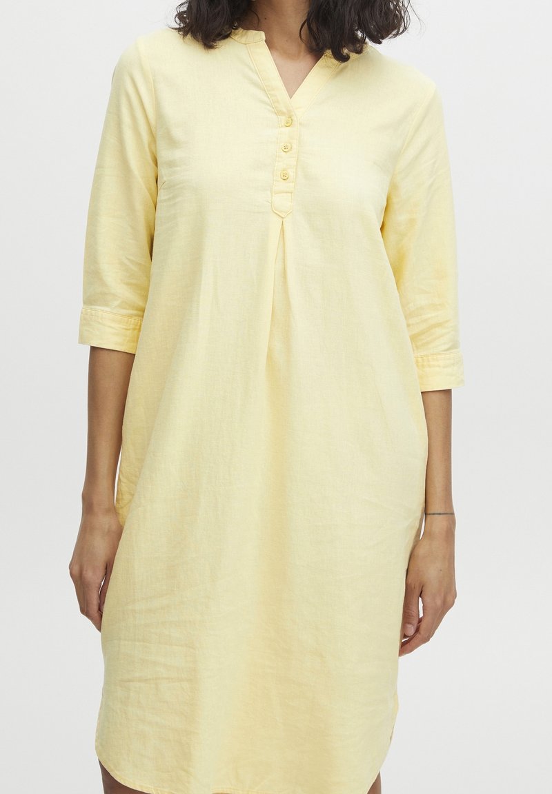 Femme portant une robe jaune pâle longueur genoux avec manches trois-quarts et un col en V boutonné sur un fond blanc uni.