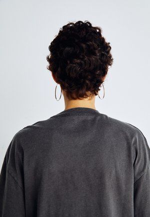 Sudadera gris de gran tamaño con superficie texturizada y hombros caídos, vista desde atrás, con cabello corto y rizado y pendientes de aro.