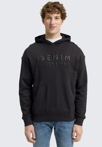 Crna hoodie s natpisom "DENIM DESIGN STUDIO" u izbočenom tekstu na prednjoj strani. Ima kengursku džepu, kapuljaču s vezicom i rebraste manžete.