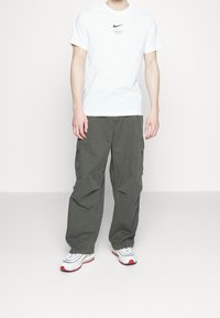 Vit Nike T-shirt med svart logga, parat med lösa olivgröna cargo-byxor. Skorna är vita med röda accenter och grå detaljer.