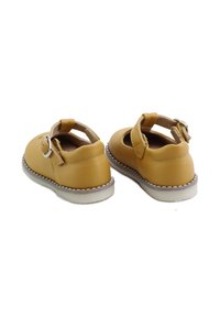 Par de zapatos para niño pequeño de color mostaza con correas de hebilla y costuras blancas alrededor de la suela, vistos desde atrás sobre un fondo blanco.