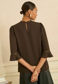 Barna blúz puffos ujjakkal és kulcslyuk hátrésszel. Az ujjak díszítő szegélyt tartalmaznak, és az anyaga sima, könnyű textúrával rendelkezik.