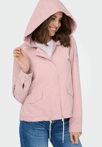 Femme souriante portant une veste à capuche rose clair avec des poches et des cordons, associée à un jean bleu, sur un fond uni.