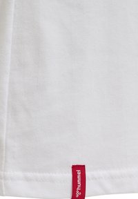 Witte stof met een rood kledinglabel genaaid bij de zoom met de merknaam "hummel" in witte tekst.