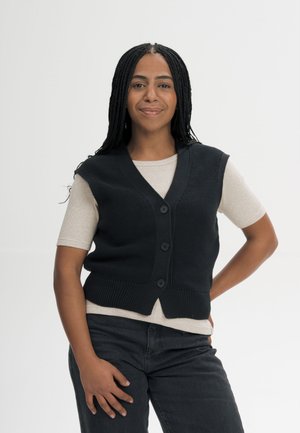 Jonge vrouw met gevlochten haar draagt een beige shirt met korte mouwen, een zwart gesloten vest en donkere broek, staand met hand op de heup tegen een witte achtergrond.