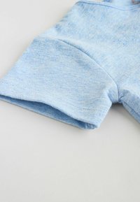 Tela de algodón azul claro, con una textura suave y puños enrollados. El diseño es informal, con un ligero patrón jaspeado visible.