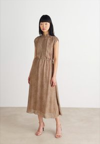 Kratkoročna, rjava obleka z vzorcem in visokim ovratnikom, z elastiko v pasu, ki seže pod kolena, v kombinaciji z natikači v bež barvi.