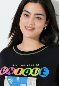Sort t-shirt med korte ærmer, med rund halsudskæring med grøn syning og et farverigt grafisk tryk med teksten "ALT DU BEHØVER ER UNIKT."