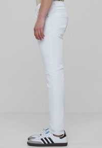 Jeans albe slim-fit cu detalii distruse la genunchi, asociate cu adidași clasici albi, cu dungi negre și accente gri.
