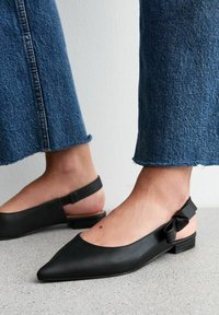 Svart läder slingback-lägenheter med spetsig tå, med en liten rosettdetalj vid hälen och låg profil. Texturerad svart yttersula.