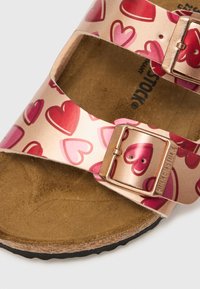 Rožnati in zlati sandali z vzorci src, dvema nastavljivima trakoma in pluto podplatom. Imajo kovinske sponke in teksturiran podplat.