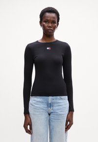 Top nero a maniche lunghe a costine con scollatura rotonda, caratterizzato da un piccolo logo colorato ricamato sul petto. Indossato con jeans blu chiaro.