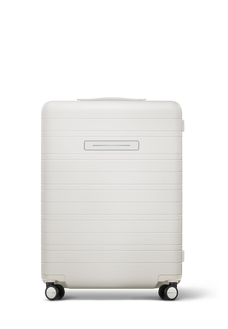 Valise rigide blanche avec des rainures horizontales, une poignée supérieure, un compartiment pour ID slim, et quatre roues noires pour la mobilité.