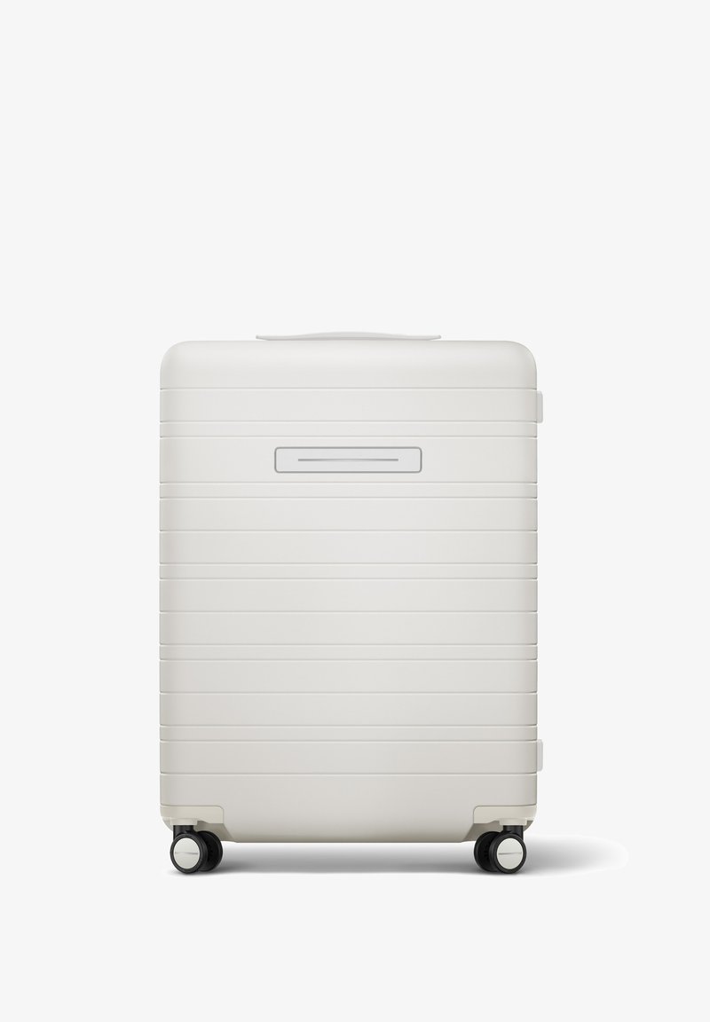 Valise rigide blanche avec des rainures horizontales, une poignée supérieure, un compartiment pour ID slim, et quatre roues noires pour la mobilité.