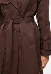 Trench-coat imperméable marron avec taille cintrée par une ceinture, boutons croisés et poches latérales, au toucher lisse.
