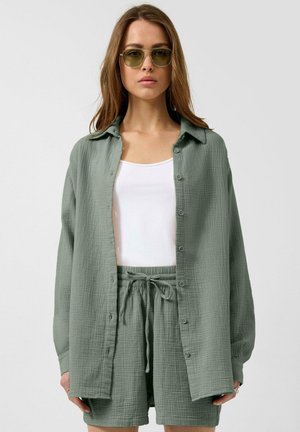 Frau trägt ein grünes, strukturiertes Hemd mit Knöpfen und passende Shorts, darunter ein weißes Tanktop und grün getönte Sonnenbrillen.