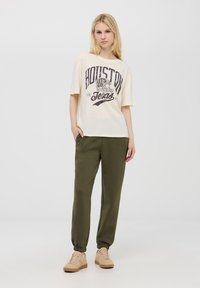 Kremowa koszulka z czarnym napisem "HOUSTON LET'S GO", połączona z oliwkowymi spodniami dresowymi i beżowymi sneakersami z jasnymi akcentami.