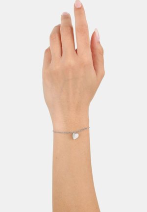 Ingrandimento di un polso che indossa un delicato braccialetto in argento con un piccolo ciondolo a forma di cuore su uno sfondo chiaro.