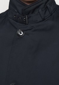 Tmavě modrá bunda s vysokým límcem, knoflíkovým zapínáním a strukturovanou tkaninou s viditelným prošitím a kulatým černým knoflíkem.