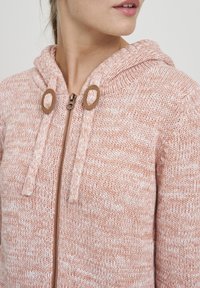 Närbild av en kvinna som bär en rosa och vit stickad zip-up huvtröja med brun dragkedja och snörningsöglor.