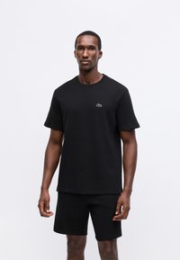 Homem em pé, vestindo uma camisa preta texturizada de mangas curtas com um pequeno logótipo de crocodilo e calções pretos a condizer, sobre um fundo branco liso.