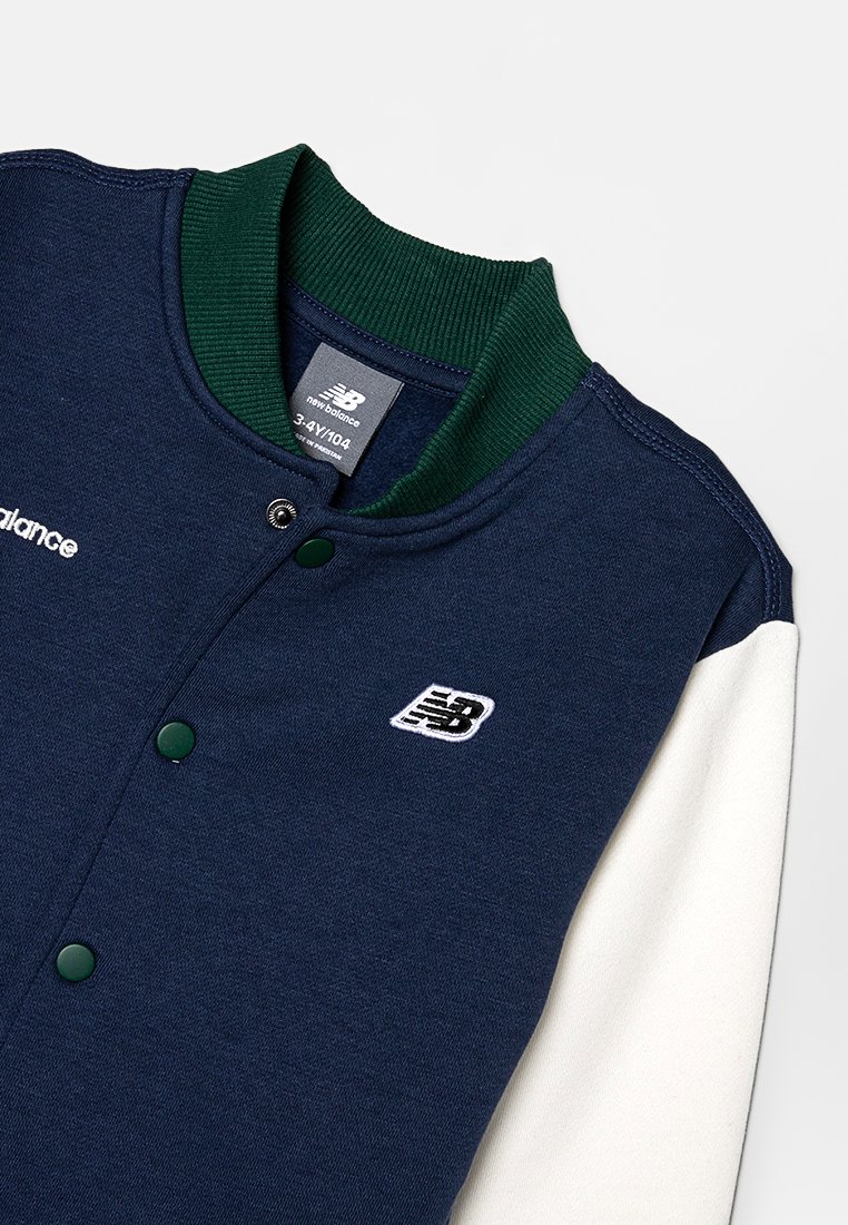 Veste de style varsity bleu marine avec col côtelé vert, manches blanches, boutons-pression et logo brodé. Texture douce, design décontracté.