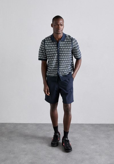 Chemise à manches courtes, à motifs marins avec des motifs en zigzag, associée à un short sombre et des baskets noires, sur un fond gris.