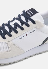 Fehér sneaker struktúrált velúrból és hálós anyagból készült felsőrésszel, tengerészkék fűzőkkel és fekete logócimkével, amelyen a "Tommy Hilfiger" felirat szerepel. Szürke gumitalp.