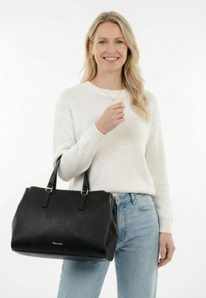 Mujer sonriente con suéter blanco y jeans azul claro sostiene un gran bolso de cuero negro con un logo dorado.