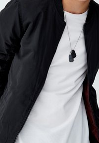 Blouson aviateur noir avec une fermeture éclair, des poignets côtelés et une doublure bordeaux, porté sur un t-shirt blanc uni. Comprend une chaîne argentée avec des plaques d'identification.