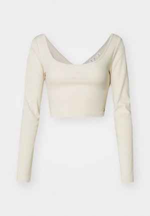 Dlhý rukáv cropped top v svetlej béžovej rebrovanej látke. Obsahuje široký výstrih a bočný akcent s gombíkom. Hladká textúra.