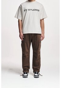 T-shirt en coton écrème avec le texte noir "2Y STUDIOS", associé à un pantalon cargo marron doté de poches et d'ourlets à cordon, porté avec des baskets noires.