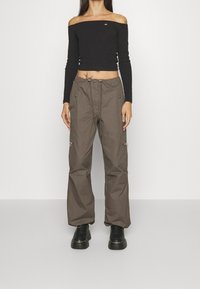 Schwarzes schulterfreies Oberteil mit langen Ärmeln, kombiniert mit lockeren braunen Cargo-Hosen mit Taschen und metallischen Reißverschlüssen. Gestylt mit schwarzen Plateau-Stiefeln.