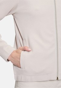Veste texturée beige clair avec fermeture éclair à l'avant, poches latérales et poignets côtelés. Le tissu présente un motif tissé subtil.