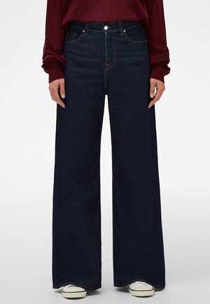 Osoba ubrana w ciemnoniebieskie szerokie jeansy, bordową bluzkę z długim rękawem i białe trampki, stojąca na jednolitym tle.