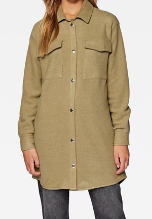 Lange olijfgroene jas met knopen en twee klepzakken op de borst, gedragen over donkergrijze jeans.