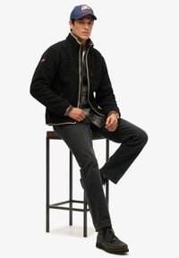 Zwarte fleecejas met ritssluiting, gestructureerd oppervlak, crème bies, gedragen over een geruit flanellen overhemd. Donkere spijkerbroek en zwarte laarzen maken de look compleet.