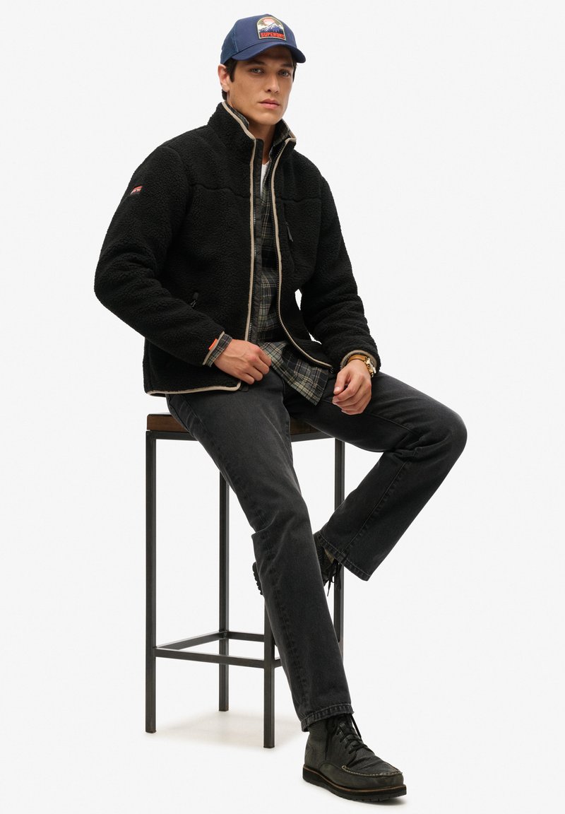 Zwarte fleecejas met ritssluiting, gestructureerd oppervlak, crème bies, gedragen over een geruit flanellen overhemd. Donkere spijkerbroek en zwarte laarzen maken de look compleet.