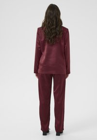 Donna con lunghi capelli ondulati che indossa un completo di satin bordeaux e tacchi neri, in piedi con la schiena a una semplice sfondo chiaro.